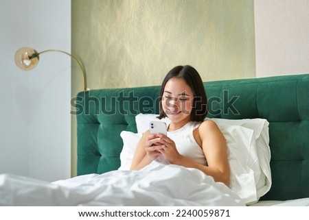 Cute korean girl in bed, holding smartphone, feeling happy and pleased, spending morning in bed, enjoying surfing net on mobile phone.