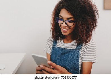 Cute Lady With Bronze Skin Checking New Messages In Social Networks Using Her Phone. Indoor Portrait Of Smiling Curly Girl Testing Mobile App On Smartphone Sitting On Light Background.