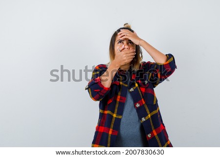  cute lady keeping hands on mouth and forehead, looking up in checked shirt and looking wondered , front view. 