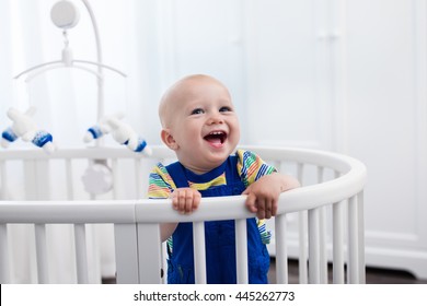 Cute laughing baby standing in a white round bed. White nursery for young children. Little boy learning to stand in his crib. Toys for infant cot. Smiling child playing with toy bear in sunny bedroom.
