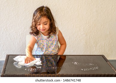 Cute Laughing Curly Toddler Girl Washing Glass Table With Paper Towel In Home Terrace, With Water And Detergent Solution Spray.