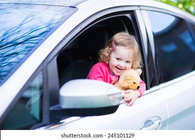 Cute Laughing Curly Toddler Girl Playing With Toy Teddy Bear Sitting In A Silver Color Modern Family Car On Front Seat Watching Out A Window In A Side Mirror Enjoying Weekend Vacation Ride After Rain 