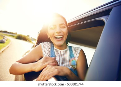 Cute Laughing Young Woman Hanging Out Her Head From A Car Through The Open Window Enjoying The Freedom Of The Breeze In Her Hair As It Travels Along A Rural Road