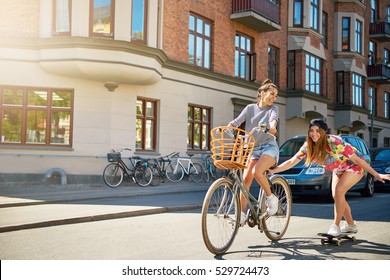 Cute Laughing Young Woman In Jeans Shorts On Bike With Basket Pulling Girl In Tie Die Shirt And Black Hat Holding On While Using Skateboard