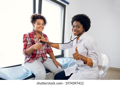 Cute Likable Mixed Race Teen Girl And Joyful African Woman Doctor, Showing Thumbs Up During Doctors Checkup. Pediatrist Examinate Young Patient's Heartbeat And Lungs With Stethoscope