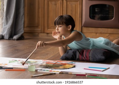 Cute Little 8s Latino Girl Kid Lying On Floor At Home Have Fun Drawing With Colors In Album On Weekend. Happy Small Hispanic Child Engaged In Painting Artistic Activity At Home. Hobby, Art Concept.