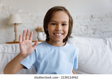 Cute little 9s boy sit on couch at home wave hand start video call conversation with tutor or friend, communicates remotely smile look at camera. Virtual meeting, young gen using modern tech concept