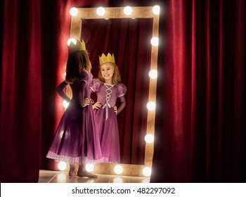 Cute Little Actress. Child Girl In Princess Costume On The Background Of Theatrical Scenes And Mirrors.