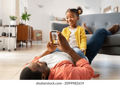 Cute Little African American Girl Sitting On Father's Stomach And Playing With Him. Mature Black Father And Daughter Enjoying At Home By Clicking Pictures In Mobile Phone. Lovely Dad Taking Picture.