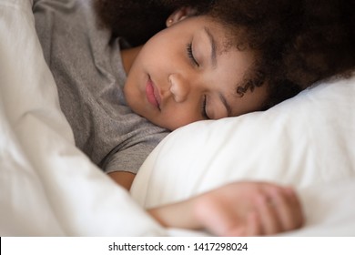 Cute little african american kid girl sleeping well alone in bed under warm blanket duvet lying on comfort white soft pillow, adorable small child rest asleep enjoy good healthy peaceful sleep or nap