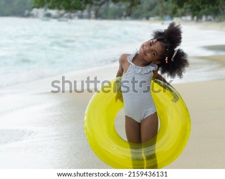 Cute little African girl posing for camera on the beach. Little girl wearing blue stripy swimming suit holding neon yellow transparent swim ring. Summer holidays concept;.