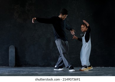 Cute Little African Kid Boy And Cool Young Asian Man Teacher With Tattoo Are Practice Hip Hop Or Freestyle Dancing On Cement Floor On Black Background. Child Education On Free Time