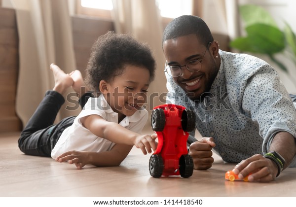 かわいいアフリカの小さな息子が 黒い父親とおもちゃの車を遊び 幸せな家族の小さな混血児と 家で暖かい床で楽しいレースをする若い父親 のベビーシッター 家族の父親の子どもの遊び の写真素材 今すぐ編集