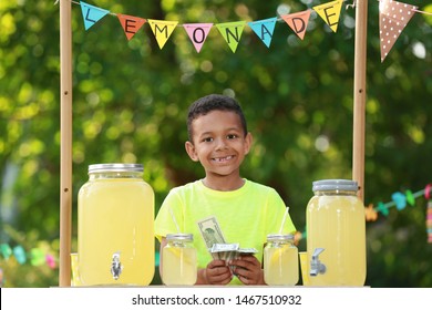 Cute Little African-American Boy With Money At Lemonade Stand In Park. Summer Refreshing Natural Drink