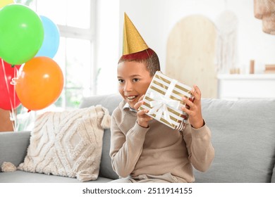 Cute little African-American boy in party hat with gift box celebrating birthday and sitting on sofa in living room - Powered by Shutterstock