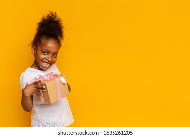Cute little afro girl smiling and holding gift box over yellow background, copy space