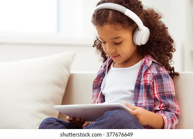 Cute little Afro-American girl in casual clothes and headphones using a tablet and listening to music while sitting on a sofa in the room. - Powered by Shutterstock