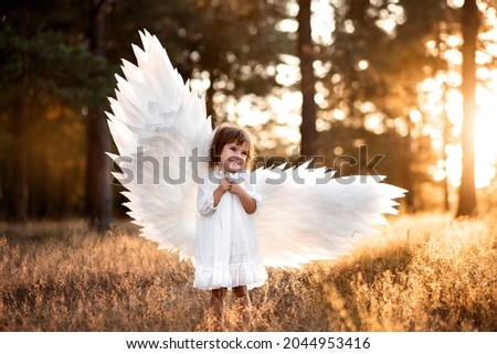 Cute little angel girl in white clothes, dress and wings, standing where the light of sunset, the sun's rays in the summer forest. Gentle, artistic image, photo, glare.