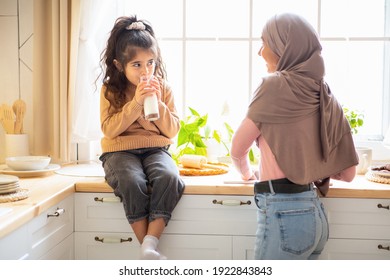 Cute Little Arab Girl And Her Muslim Mom In Hijab Spending Time Together In Kitchen, Small Female Kid Sitting At Table And Drinking Milk From Glass While Islamic Mother Washing Dishes, Free Space