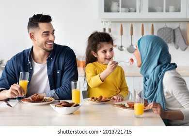 Cute Little Arab Girl Playfully Feeding Mom While Having Breakfast With Parents In Kitchen, Happy Muslim Family Of Three Sitting At Table And Enjoying Tasty Morning Meal Together, Closeup Shot