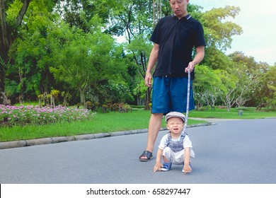 Cute Little Asian 1 Year Old Toddler Baby Boy Child In Beautiful Park Tethered On A Leash And Walking Along A Pavement, Dad And Son Cross Over The Road In Public Park, Kid Strapped To A Backpack Leash