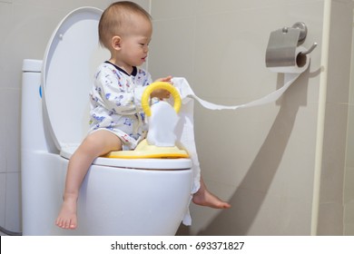 Cute Little Asian 1 Year Old Toddler Baby Boy Child Sitting On The Toilet Modern Style With A Kid Bathroom Accessory And Holding & Playing With Toilet Paper. Potty / Toilet Training Child Concept.