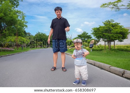 Cute little Asian 1 year old toddler baby boy child in beautiful park tethered on a leash and walking along a pavement, Dad and son cross over the road in public park, kid Strapped to a Backpack Leash