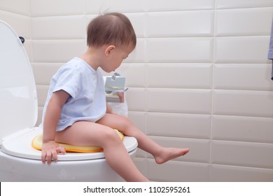 Cute Little Asian 2 Year Old Toddler Baby Boy Child Sitting On The Toilet Modern Style With A Kid Bathroom Accessory And Holding & Playing With Toilet Paper. Potty / Toilet Training Child Concept