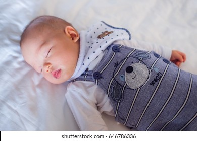 Cute Little Asian 5 Months Old  Baby Boy Child In Long Dungarees And White T-shirt Costume Wearing Bib Sleeping Mouth Open On White Sheet On Bed In Bedroom At Home