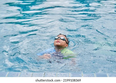 Cute Little Asian 6 Years Old School Boy Child In Swimming Goggles Learning To Swim, Floating, Treading Water To Stay Afloat At Outdoor Pool In Summer Day, Happy Kid Having Fun On Summer Vacation