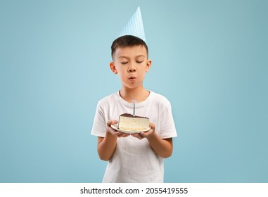 Cute Little Asian Boy In Party Hat Blowing Candle On Birthday Cake, Happy Small Male Child Holding Piece Of Sweet B-Day Treat, Making Wish While Standing Over Blue Studio Background, Copy Space