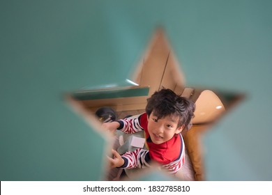 Cute Little Asian Boy Smile And Look Up Through A Star Shaped Hole On Pastel Vintage Blue Cardboard Wall At The Camera, While Sitting In Cardboard House In Living Room At Home.