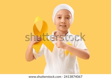 Cute little Asian girl after chemotherapy pointing at yellow ribbon on color background. Childhood cancer awareness concept