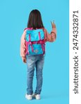 Cute little Asian girl with backpack showing victory gesture on blue background, back view. End of school year