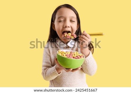 Cute little Asian girl eating cereal rings on yellow background