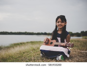 Cute little asian girl sitting on grass and play ukulele in park.