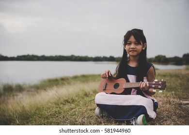 Cute little asian girl sitting on grass and play ukulele in park.