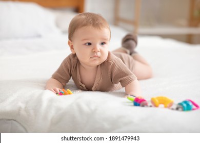 Cute Little Baby Boy Or Girl Playing With Toys While Laying On Bed At Home, Looking At Copy Space. Adorable Toddler Having Fun, Lying On Belly And Palying With Beanbag. Kids Toys, Maracas Concept