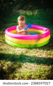 A cute little baby boy is playing with toys while bathing in a little colorful wading pool on meadow garden field outdoor. Summer time. Joy and happiness. Love and family 