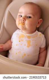 Cute Little Baby Boy Sitting In High Chair All Stained With Porridge, Making Funny Faces And Stretching While Refusing To Eat