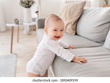 Cute Little Baby Boy Smiling To Camera Standing Near Couch In Living Room At Home. Happy Toddler Having Fun Alone. Childhood, Child Care And Parenthood Concept