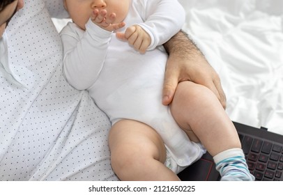 cute little baby in father's arm is playing with his hands. father and son during pandemic, working online from home. black laptop on legs. boys are sitting on the bed for kids. fatherhood concept.