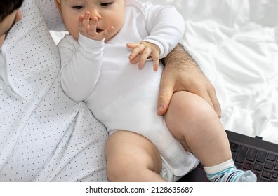 Cute Little Baby In Father's Arm Is Playing With His Hands. Father And Son During Pandemic, Working Online From Home. Black Laptop On Legs. Boys Are Sitting On The Bed For Kids. Fatherhood Concept.