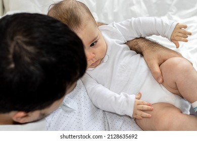 Cute Little Baby In Father's Arm Is Playing With His Hands. Father And Son During Pandemic, Working Online From Home. Black Laptop On Legs. Boys Are Sitting On The Bed For Kids. Fatherhood Concept.