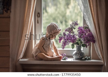 Cute little baby girl in a bonnet on the windowsill looking at beautiful lilac bouquet in a vase. Image with selective focus and toning
