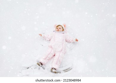 Cute little baby girl in colorful winter clothes making snow angel, laying down on snow. Active outdoors leisure with children in winter. Toddler in pink snowsuit.