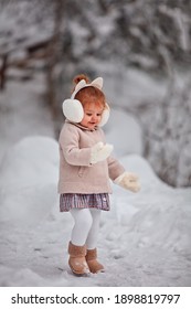 Cute Little Baby Girl Having Fun In Winter Park, Playing With The Snow, Wearing Gloves And Earmuffs