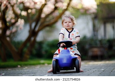 Cute Little Baby Girl Playing With Blue Small Toy Car In Garden Of Home Or Nursery. Upset Crying Toddler Child Outdoors.