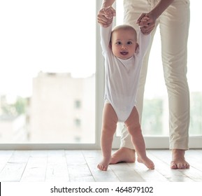 Cute little baby is looking at camera and smiling while learning to walk, mom is holding his hands