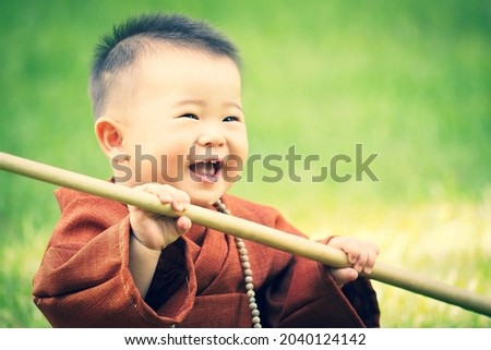 Cute little baby monk holding his wooden stick in the grass field, happy and smiling.